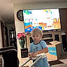 blue_shirt, blur, cabinet, cardboard_box, carpet, cartoon, child, clock, diaper, floor, flowers, furniture, home_interior, living_room, plastic_toy, television, toddler, toy_walker, vase, window