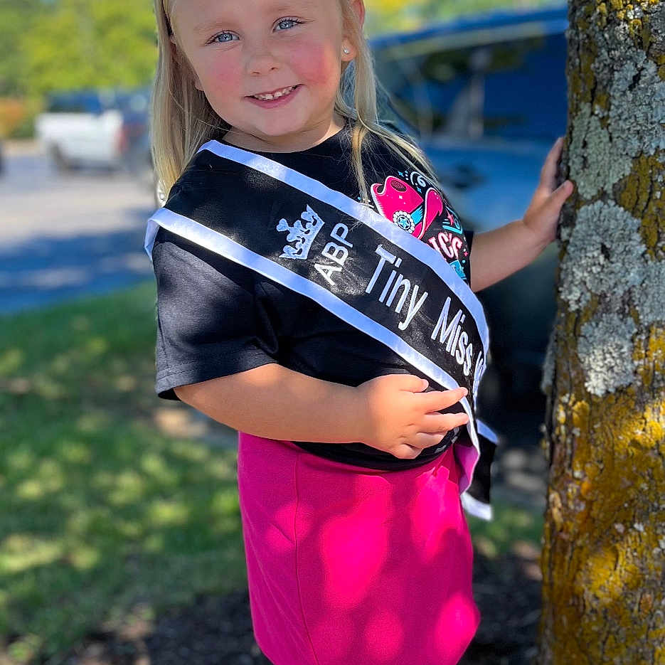Brylee is registered to the contest to win money with this photo: child, clothing, dress, face, female, footwear, girl, grass, head, nature, outdoors, person, photography, plant, portrait, shoe, shorts, skirt, standing, tree