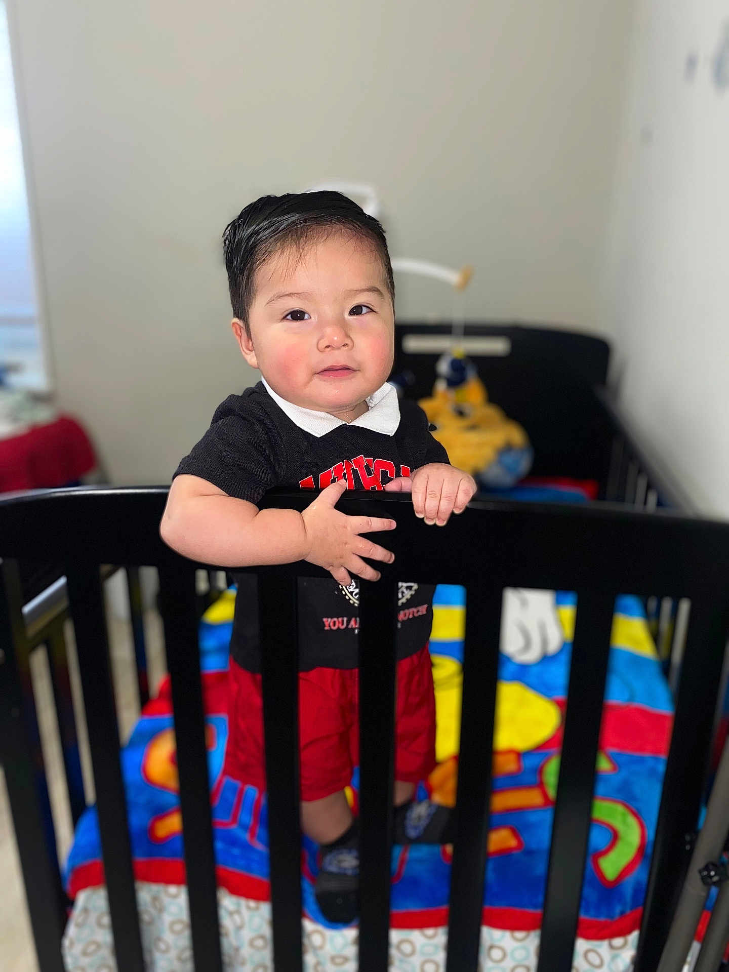Ethan is registered to the contest to win money with this photo: toddler, child, crib, black_shirt, red_shorts, colorful_blanket, toy, indoor, standing, baby, hand, face, hair, smile, person, footwear, home, furniture, blurred_background, light