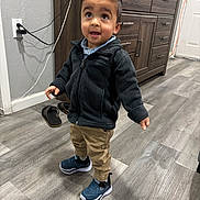 Aayansh is registered to the contest to win money with this photo: child, toddler, standing, smile, sneakers, jacket, pants, indoor, hardwood_floor, dresser, cabinet, flip_flops, charging_cable, wall_socket, portrait, hair, eyes, curiosity, flooring, room