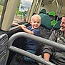 child, toddler, bus, window, seat, adult, woman, smile, holding, clothing, interior, public_transport, handrail, blue_shirt, jeans, blond_hair, patterned_seat, green_headrest, daylight, smiling