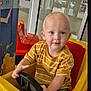 Carson is registered to the contest to win money with this photo: toddler, child, toy_car, yellow_car, steering_wheel, indoor, play_area, striped_shirt, blue_eyes, short_hair, casual_clothing, window, glass_door, colorful_mural, red_seat, jeans, cute, smiling, playing, young_child