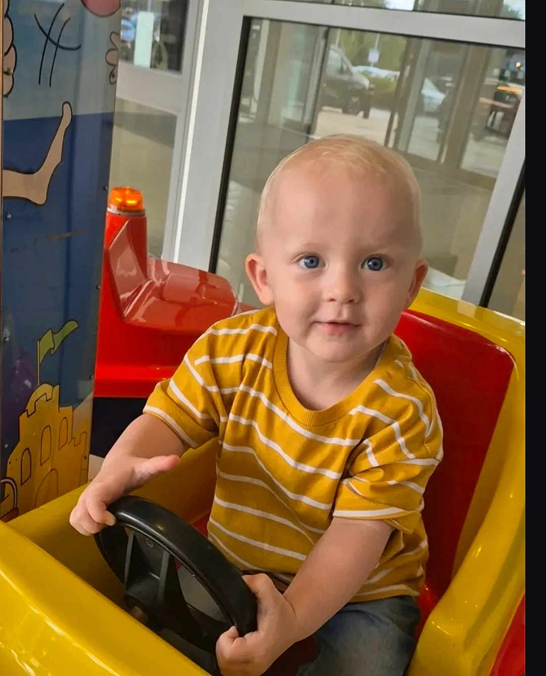 Carson is registered to the contest to win money with this photo: toddler, child, toy_car, yellow_car, steering_wheel, indoor, play_area, striped_shirt, blue_eyes, short_hair, casual_clothing, window, glass_door, colorful_mural, red_seat, jeans, cute, smiling, playing, young_child