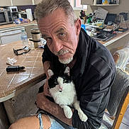 Rorschach joined the competition — help win amazing prizes! man, cat, black_and_white_cat, kitchen, chair, table, shorts, gray_hair, facial_hair, indoor, household_items, water_bottle, microwave, serious_expression, pet, holding, seated, wooden_chair, countertop, casual_clothing
