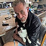 man, cat, black_and_white_cat, kitchen, chair, table, shorts, gray_hair, facial_hair, indoor, household_items, water_bottle, microwave, serious_expression, pet, holding, seated, wooden_chair, countertop, casual_clothing