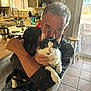 Rorschach is registered to the contest to win money with this photo: man, cat, indoor, kitchen, chair, tile_floor, window, natural_light, black_and_white_cat, affection, seated, home, casual_clothing, facial_expression, domestic_life, pet, holding, relaxed, background_objects, daylight