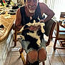 Rorschach is registered to the contest to win money with this photo: man, cat, indoor, chair, smile, beard, black_and_white_cat, wooden_chair, tank_top, shorts, tiled_floor, table, kitchen_counter, window, sunlight, casual, earbuds, relaxed, clutter, home