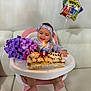 Antonella is registered to the contest to win money with this photo: baby, child, high_chair, birthday, cake, candle, flower_bouquet, purple_flowers, balloon, headband, smiling, happy, indoor, celebration, dress, foot, sofa, white_couch, table, party