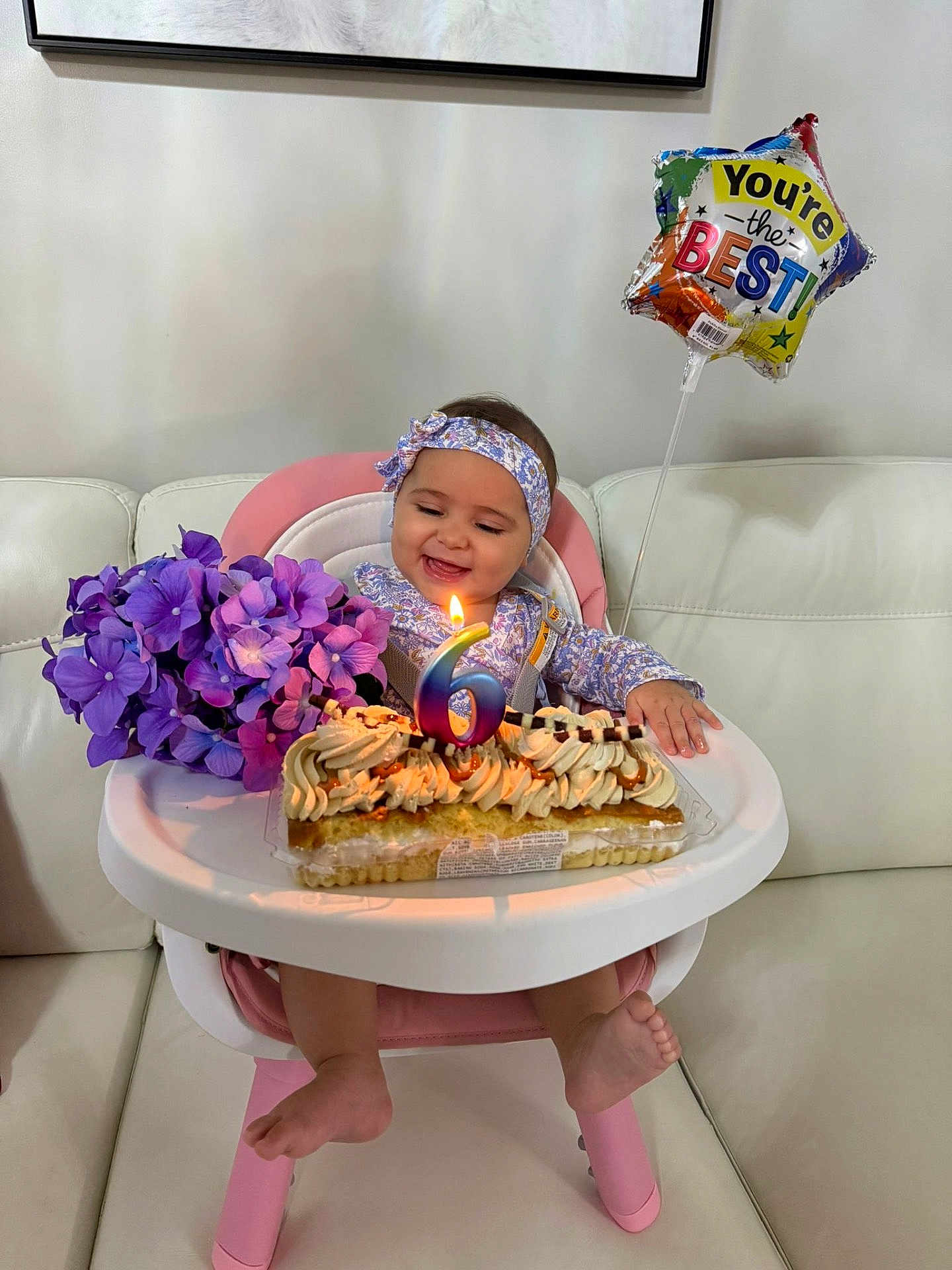 Antonella is registered to the contest to win money with this photo: baby, child, high_chair, birthday, cake, candle, flower_bouquet, purple_flowers, balloon, headband, smiling, happy, indoor, celebration, dress, foot, sofa, white_couch, table, party