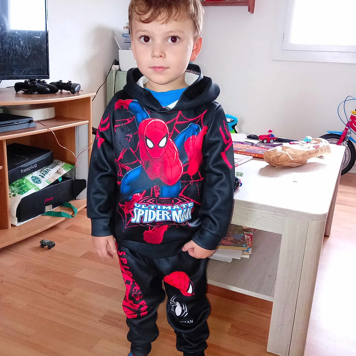 Maëlone participe au concours pour gagner de l'argent avec cette photo : books, boy, child, clothing, floor, gaming_controller, hoodie, indoor, living_room, neutral_expression, pants, person, shelf, socks, spiderman, table, toys, tv, window, wooden_floor