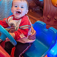 Freddie joined the competition — help win amazing prizes! adult_hands, blue_lights, blue_steering_wheel, bright_colors, casual_clothing, child, face, fun, happy, indoor, joy, person, play_area, play_car, red_clothing, seat, short_hair, smiling, toddler, toy