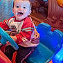 adult_hands, blue_lights, blue_steering_wheel, bright_colors, casual_clothing, child, face, fun, happy, indoor, joy, person, play_area, play_car, red_clothing, seat, short_hair, smiling, toddler, toy