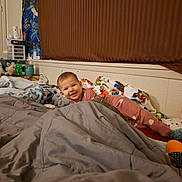 Juliana is registered to the contest to win money with this photo: baby, child, smiling, bedroom, bed, blanket, bedding, pajamas, pink_clothing, toy, stuffed_animal, nursery, indoor, portrait, happy, lying_down, cozy, window_blinds, bedside_table, household_items