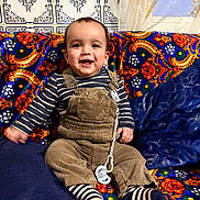Soulaymane a rejoint le concours — aidez-le/la à gagner de superbes lots ! baby, child, smiling, overalls, striped_clothing, socks, pacifier, couch, colorful, patterned, happy, indoors, portrait, sitting, face, person, fabric, cushion, home, cute