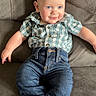 baby, child, couch, plaid_shirt, blue_jeans, socks, smile, indoor, face, person, seated, furniture, cute, young_child, relaxed, casual_clothing, blue_eyes, bald, happy, infant