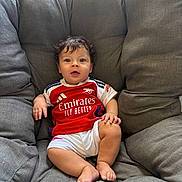 Mohamed joined the competition — help win amazing prizes! baby, child, curly_hair, red_jersey, white_shorts, sitting, grey_chair, cushion, indoors, comfortable, cute, portrait, smiling, face, person, young_child, casual, relaxed, sportswear, happy