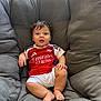 baby, child, curly_hair, red_jersey, white_shorts, sitting, grey_chair, cushion, indoors, comfortable, cute, portrait, smiling, face, person, young_child, casual, relaxed, sportswear, happy