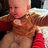 baby, child, smiling, indoor, person, clothing, red_sleeve, brown_top, beige_pants, fluffy_surface, hand, arm, happy, cute, infant, sitting, face, head, skin, cozy