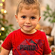 Kayden is registered to the contest to win money with this photo: child, toddler, portrait, face, smiling, looking_at_camera, red_shirt, puma_shirt, topknot, brown_hair, young_person, indoor, warm_lighting, bokeh, string_lights, plant, couch, cozy, close_up, fashion