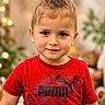 child, toddler, portrait, face, smiling, looking_at_camera, red_shirt, puma_shirt, topknot, brown_hair, young_person, indoor, warm_lighting, bokeh, string_lights, plant, couch, cozy, close_up, fashion
