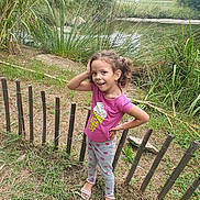 Matilde Filipa participe au concours pour gagner de l'argent avec cette photo : casual_clothing, child, curly_hair, daylight, fence, girl, grass, greenery, happy, nature, outdoor, pink_shirt, playful, polka_dot_pants, pond, sandals, smile, standing, trees, young