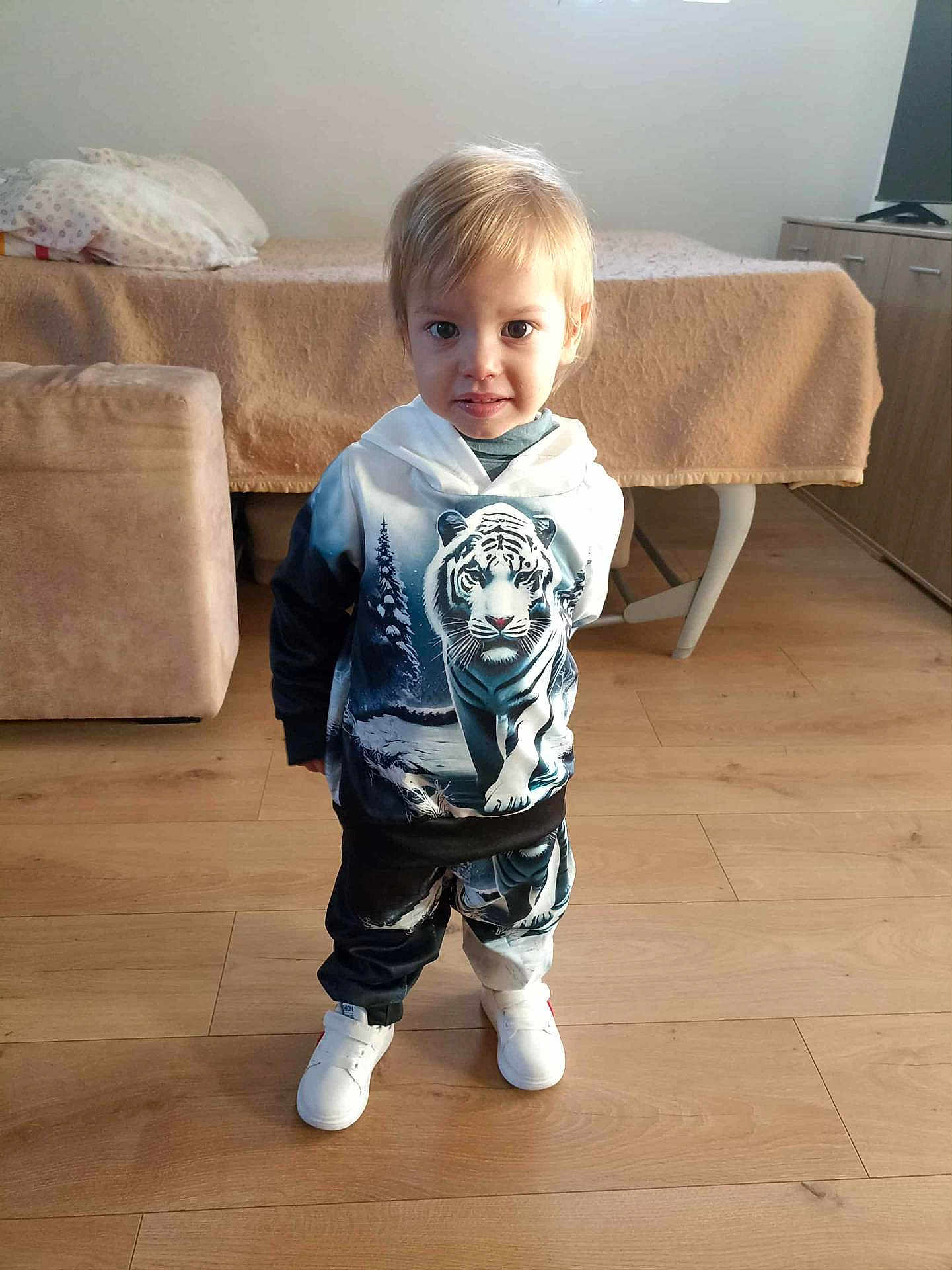 Santiago Loureiro a rejoint le concours — aidez-le/la à gagner de superbes lots ! toddler, child, hoodie, tiger_print, pants, white_shoes, wooden_floor, indoor, room, couch, bed, smiling, standing, person, clothing, furniture, light, face, hair, floor