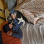 cat, person, bed, blanket, sleep_mask, pillow, indoor, resting, cozy, animal, pet, comfort, gray_cat, white_cat, hand, fabric, striped_sheet, orange_pillow, casual_clothing, relaxation