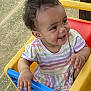 Star is registered to the contest to win money with this photo: toddler, child, smiling, curly_hair, rainbow_striped_clothing, toy_cart, yellow, blue, outdoor, grass, mesh_screen, happy, person, playtime, cute, baby, sitting, colorful, daylight, portrait