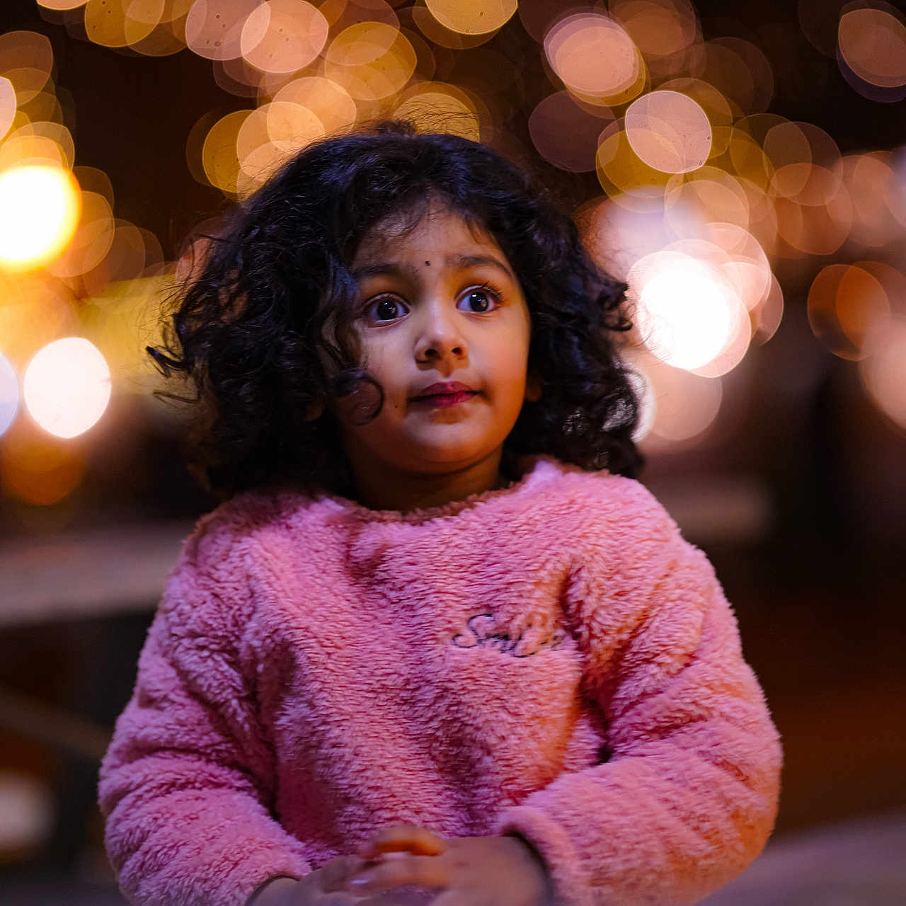 Athmiga is registered to the contest to win money with this photo: bokeh, child, cozy, curly_hair, cute, expression, face, hands, indoor, innocent, lights, looking, night, person, pink_sweater, portrait, soft_texture, warm_colors, winter_clothing, young