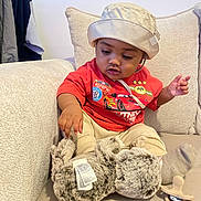Raheem participe au concours pour gagner de l'argent avec cette photo : baby, toddler, child, hat, bucket_hat, red_tshirt, animated_character, stuffed_animal, plush_toy, pacifier, couch, pillows, indoor, curious, sitting, clothing, person, face, hand, home