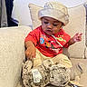 baby, toddler, child, hat, bucket_hat, red_tshirt, animated_character, stuffed_animal, plush_toy, pacifier, couch, pillows, indoor, curious, sitting, clothing, person, face, hand, home