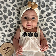 Leilani is registered to the contest to win money with this photo: baby, infant, child, headband, flower, white_top, black_pants, leopard_print_blanket, milestone_marker, wooden_plaque, cute, smiling, lying_down, portrait, indoors, soft_texture, hand, face, eyes, skin