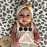 baby, infant, child, headband, flower, white_top, black_pants, leopard_print_blanket, milestone_marker, wooden_plaque, cute, smiling, lying_down, portrait, indoors, soft_texture, hand, face, eyes, skin
