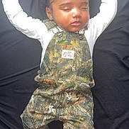 Chozyn is registered to the contest to win money with this photo: baby, sleeping, camo_overalls, white_shoes, bows, curly_hair, peaceful, infant, child, black_background, cute, adorable, fashion, footwear, hands_up, portrait, young_child, indoor, resting, clothing