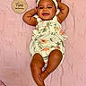 arms_up, baby, child, cute, embroidery, face, floral, happy, headband, indoors, infant, legs_crossed, lying_down, person, pink_blanket, portrait, romper, skin, smiling, wooden_sign