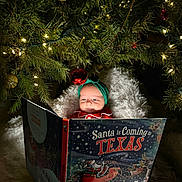 Elodie is registered to the contest to win money with this photo: baby, child, christmas_tree, holiday, book, reading, headband, blanket, lights, ornament, festive, cozy, indoor, smiling, cute, portrait, celebration, seasonal, decorations, toddler