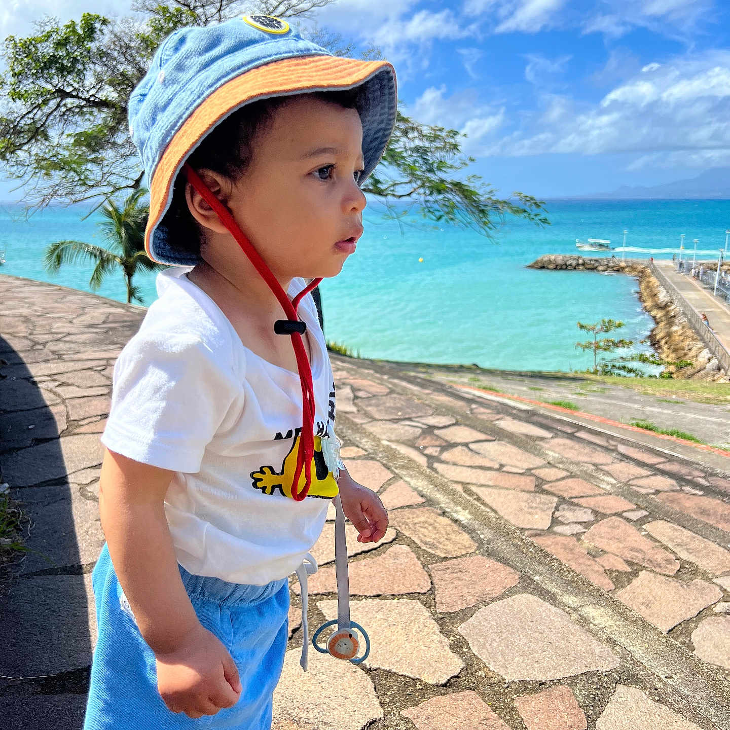 Eugène a rejoint le concours — aidez-le/la à gagner de superbes lots ! toddler, child, hat, sun_hat, toy, stethoscope, blue_sky, clouds, sea, water, stone_path, outdoor, summer, casual_clothing, sandals, greenery, tree, nature, daylight, portrait