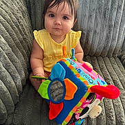 Mavis is registered to the contest to win money with this photo: baby, child, toy, colorful, soft_toy, couch, plush, yellow_clothing, indoor, curly_hair, cute, sitting, portrait, face, person, infant, playful, furniture, hand, bright