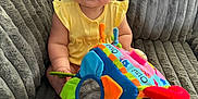 Mavis is registered to the contest to win money with this photo: baby, bright, child, colorful, couch, curly_hair, cute, face, furniture, hand, indoor, infant, person, playful, plush, portrait, sitting, soft_toy, toy, yellow_clothing