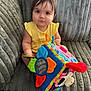 baby, child, toy, colorful, soft_toy, couch, plush, yellow_clothing, indoor, curly_hair, cute, sitting, portrait, face, person, infant, playful, furniture, hand, bright