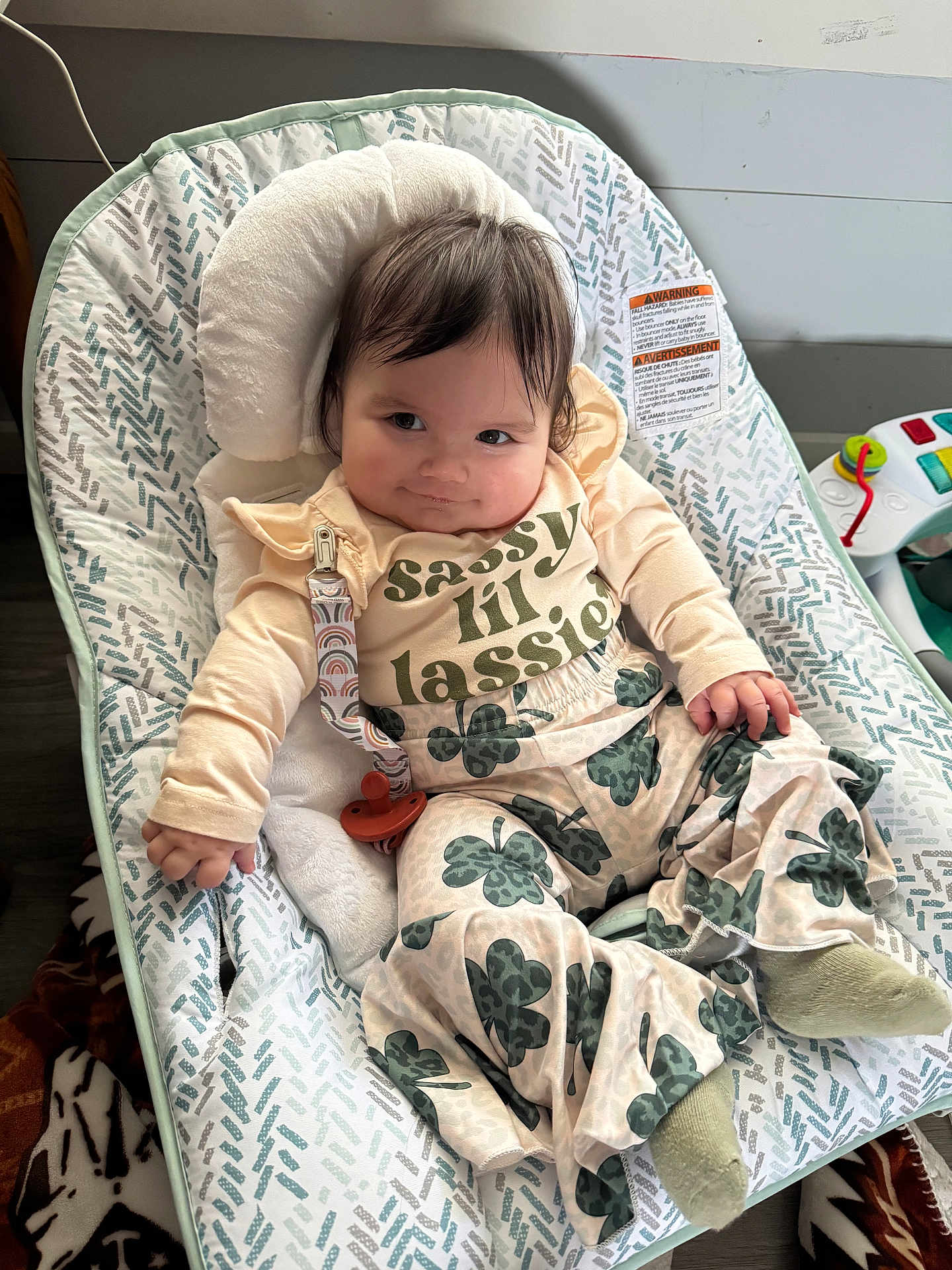 Mavis is registered to the contest to win money with this photo: baby, infant, child, baby_seat, clothing, pacifier, patterned_pants, smiling, cute, indoor, soft_toys, floor, blanket, hair, face, person, sitting, expression, comfort, playful