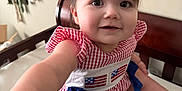 Mavis is registered to the contest to win money with this photo: baby, child, infant, dress, red, blue, gingham, crib, mattress, smile, face, arm, foot, macrame, wall_hanging, indoors, cute, person, skin, young