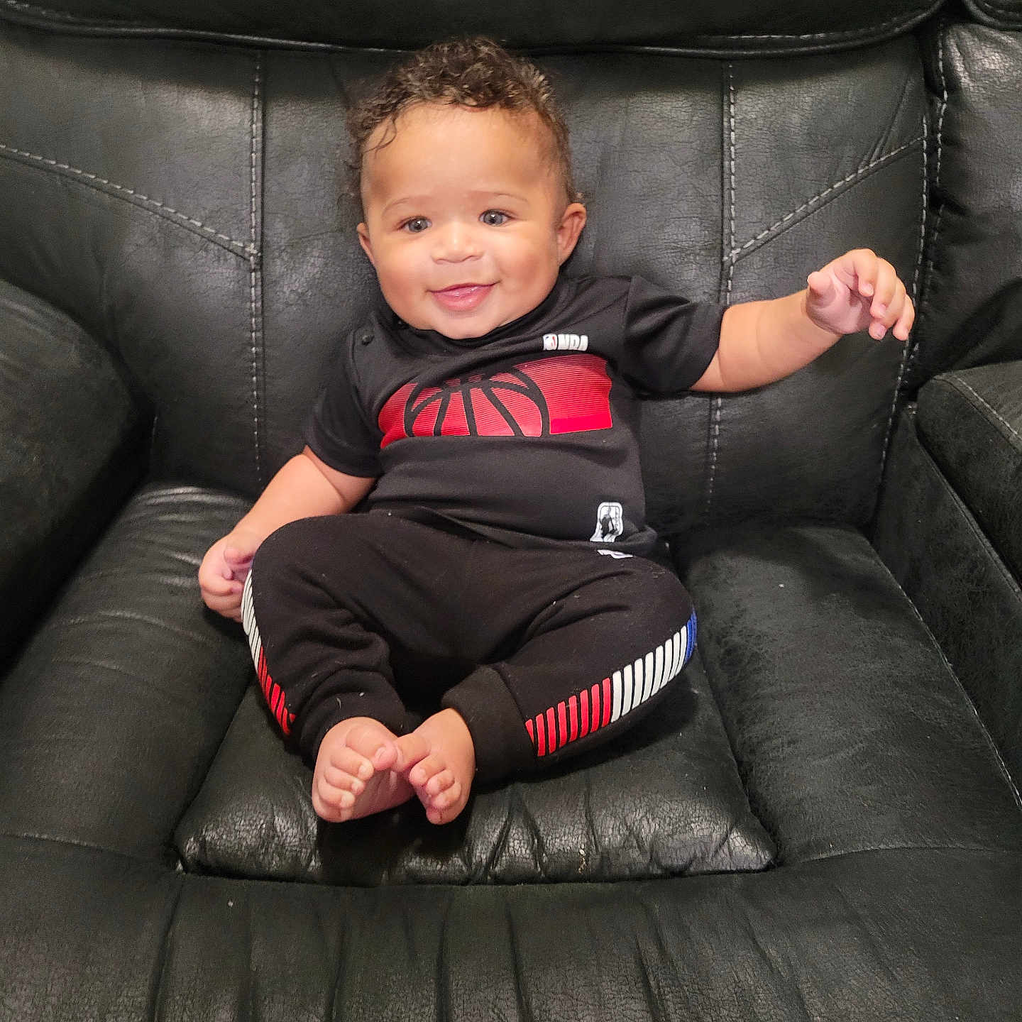 Emir is registered to the contest to win money with this photo: baby, barefoot, black_clothing, black_leather_chair, casual, child, comfortable, curly_hair, cute, furniture, happy, indoor, person, portrait, red_accents, sitting, small_toes, smiling, toddler, white_accents