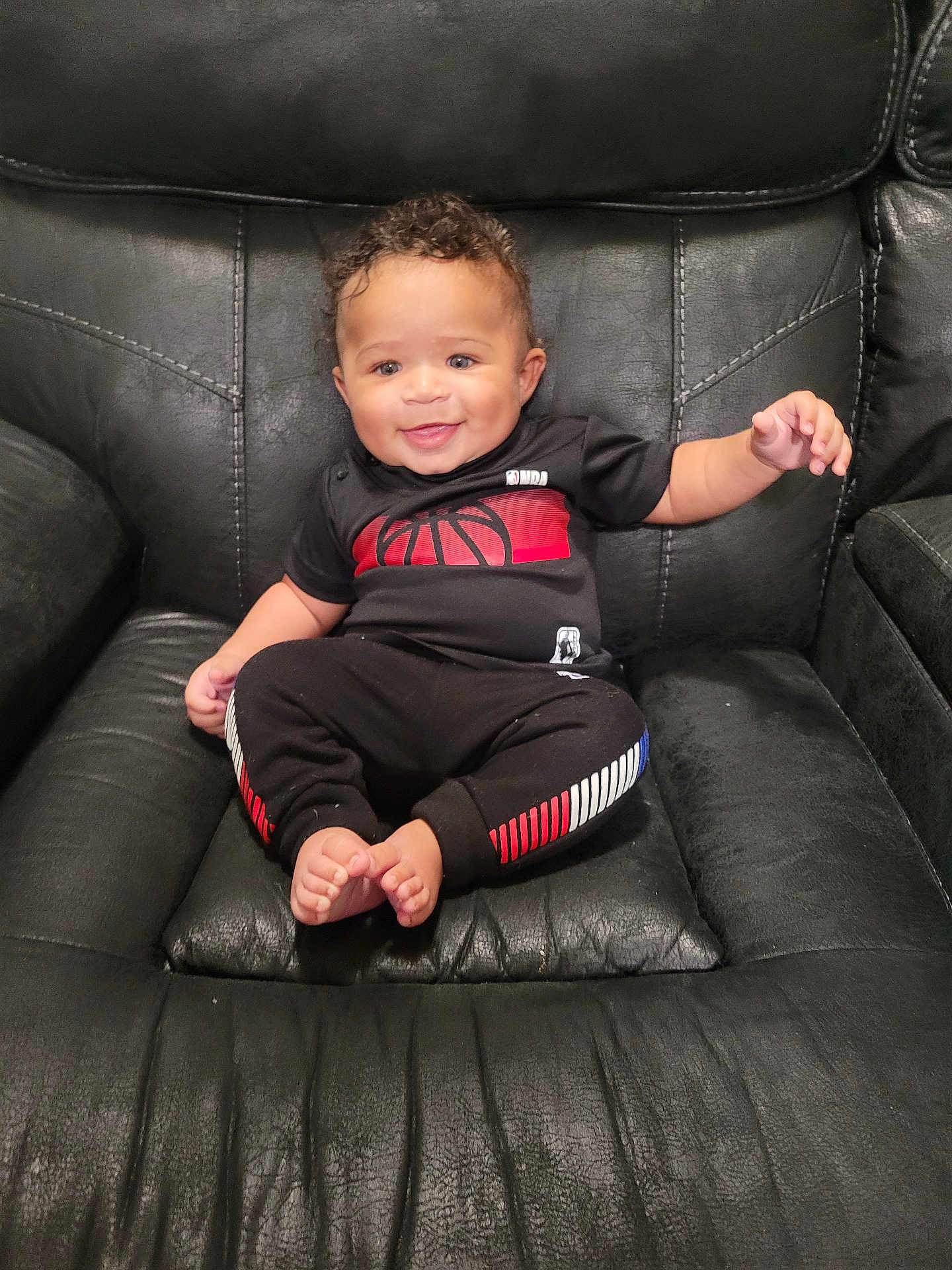 Emir is registered to the contest to win money with this photo: baby, child, smiling, black_leather_chair, curly_hair, black_clothing, red_accents, white_accents, sitting, indoor, cute, happy, portrait, toddler, barefoot, small_toes, comfortable, furniture, person, casual