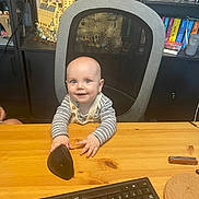 Oliver is registered to the contest to win money with this photo: baby, child, desk, computer_mouse, keyboard, office_chair, lego, bookshelf, books, games, toy, indoor, curious, smiling, person, table, furniture, striped_clothing, overalls, home_office
