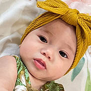 Celia is registered to the contest to win money with this photo: baby, infant, child, portrait, close_up, face, eyes, smiling, lying_down, headband, bow, mustard, green_outfit, patterned_clothing, floral_blanket, bed, indoor, baby_fashion, cute, photography