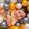 baby, baby_clothing, ball_pit, blue_eyes, child, close_up, face, gray_balls, hair, hands, happy, infant, onesie, plastic_balls, playful, portrait, smiling, toy, white_balls, yellow_balls