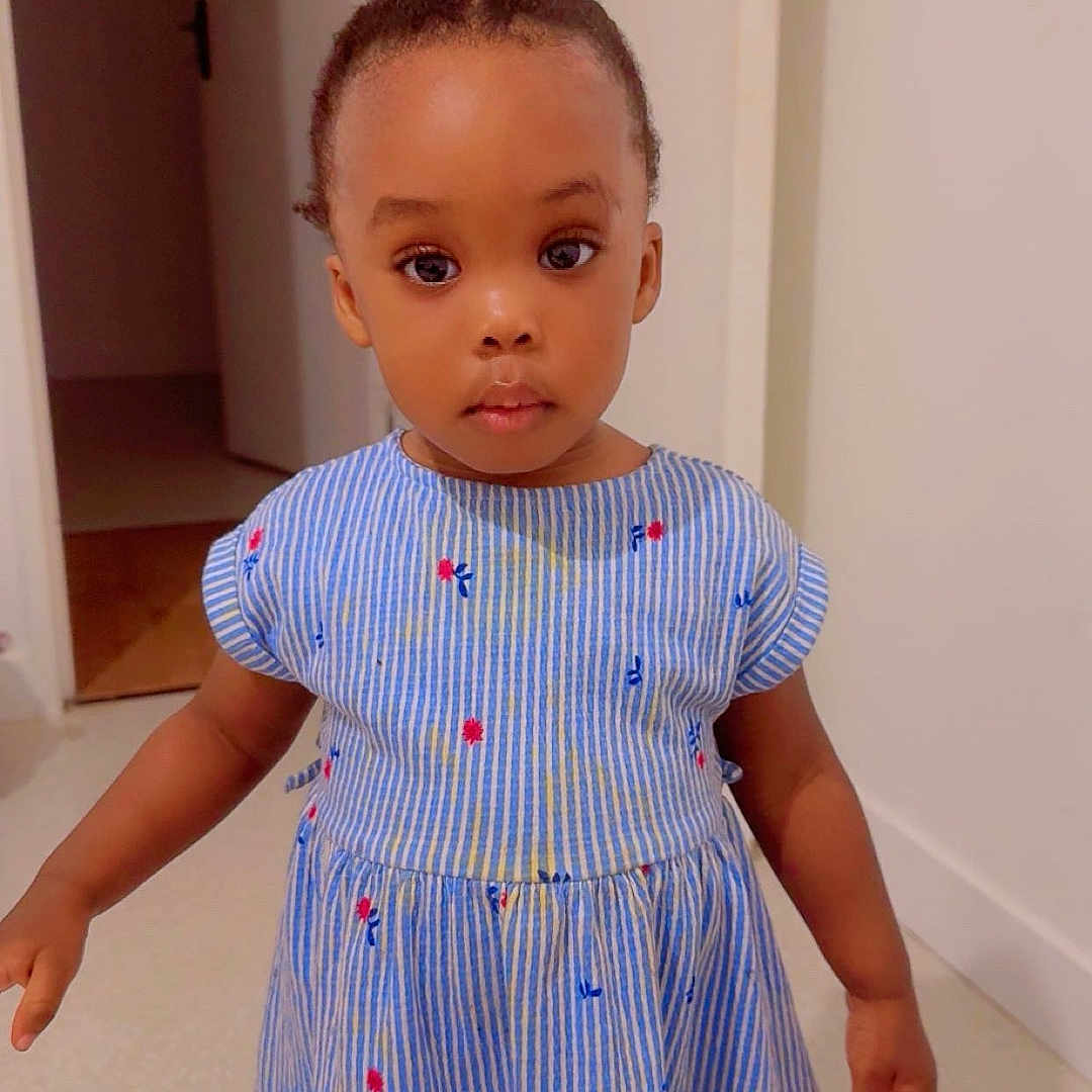 Siki Nelsia a rejoint le concours — aidez-le/la à gagner de superbes lots ! barefoot, big_eyes, blue_dress, braided_hair, carpet, child, curious_expression, door, door_handle, floral_pattern, girl, hallway, hands, indoor, photography, portrait, skin, standing, striped_dress, toddler