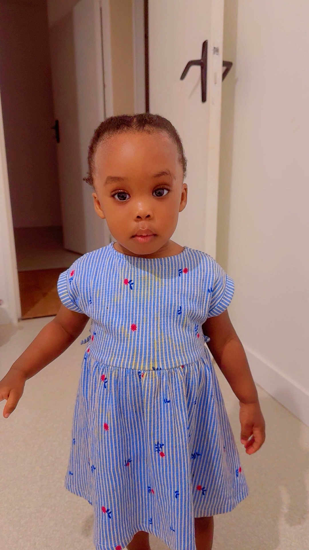 Siki Nelsia a rejoint le concours — aidez-le/la à gagner de superbes lots ! child, toddler, girl, striped_dress, blue_dress, floral_pattern, standing, hallway, door, door_handle, big_eyes, braided_hair, portrait, indoor, carpet, barefoot, curious_expression, skin, hands, photography