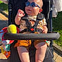 baby, stroller, sunglasses, toy, harness, buckle, legs, feet, smile, chubby_cheeks, summer, outdoors, grass, pavement, sunlit, seat, blanket, reflection, casual_clothing, plush_toy